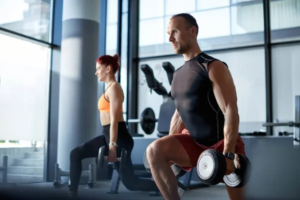 Athletic couple exercising with hand weights in lunge position during sports training in a gym. Focus is on man.