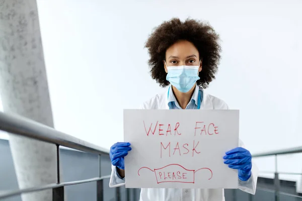 Black healthcare worker standing at medical clinic and holding placard with \'wear face  mask\' inscription due to COVID-19 pandemic.