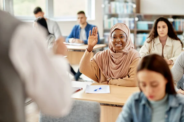 Happy black Muslim student raising her hand to answer teacher\'s question during a class in the classroom.