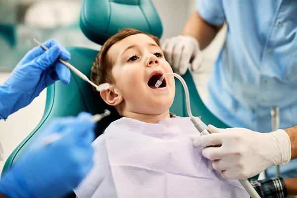 Small boy sitting in dentist\'s chair with mouth open while getting his teeth checked by dentists at dental clinic.