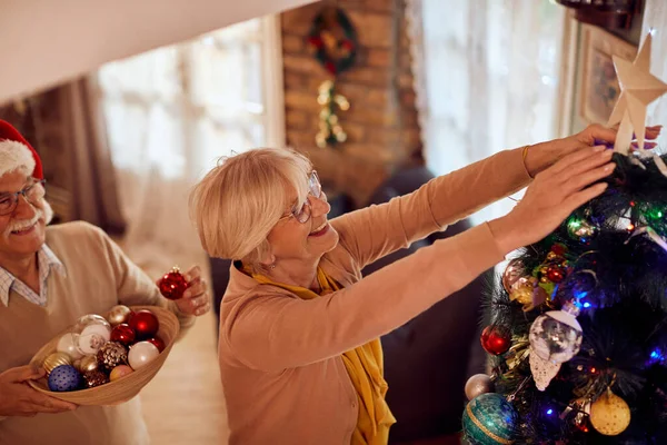 Happy mature couple having fun while decorating Christmas tree at home. Woman is putting a star on top of it.