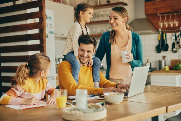 Happy parents talking to their daughters at home. Focus is on father working on laptop.