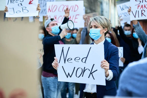 Businesswoman with protective face mask carrying banner with Need Work inscription while protesting with crowd of people on the streets.