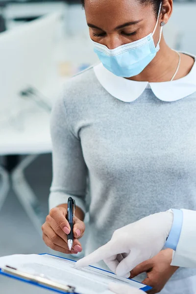 African American woman with protective face mask writing her data in medical record while having appointment with dentist at dental clinic.