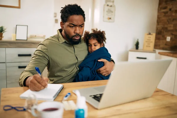 Black single father taking face of his ill daughter while working on laptop at home.