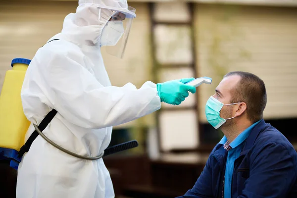 Male doctor in protective suit measuring temperature of a man with infrared thermometer during coronavirus pandemic.