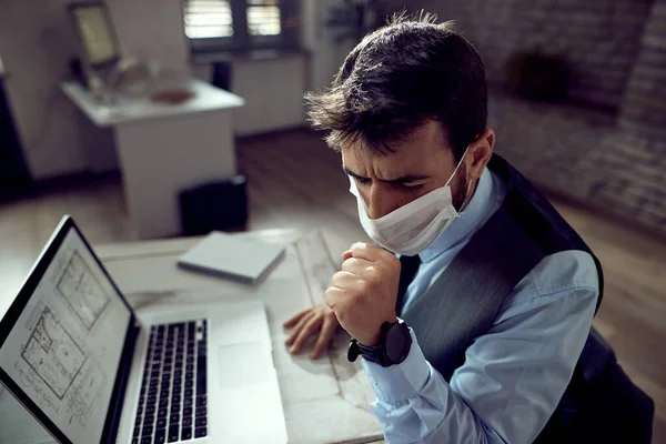 Sick businessman wearing protective face mask and coughing while working on laptop in the office.