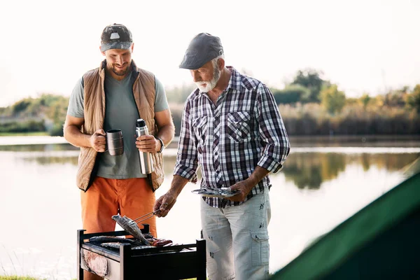 Happy man and his mature father preparing food on barbecue grill after fishing during their camping day.