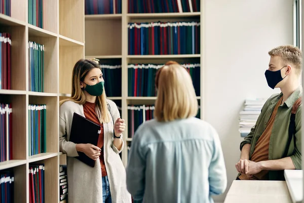 Group of college friends with protective face masks talking while studying in a library.