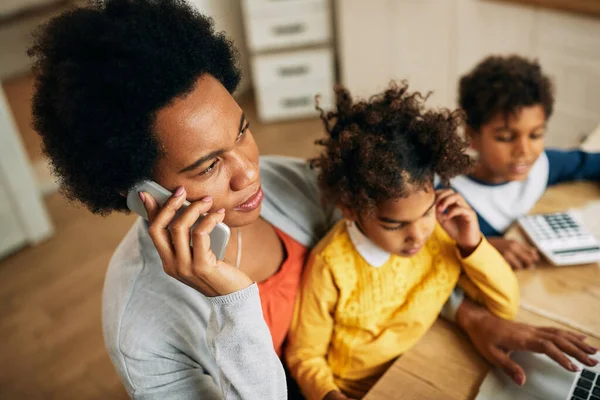 African American working mother with two children using laptop while making a phone call at home.