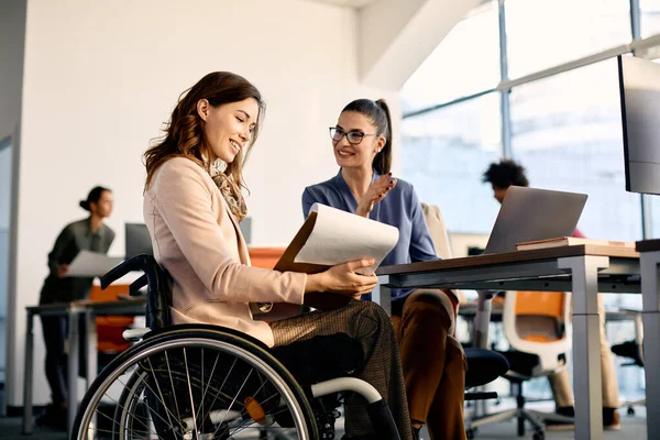Happy businesswoman in wheelchair going through reports while talking to female coworker in the office. 