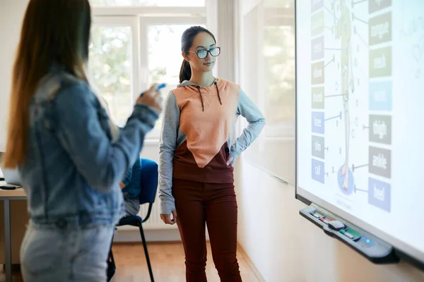 Teenager girl looking at interactive whiteboard while learning anatomy with her friend during a class at high school.