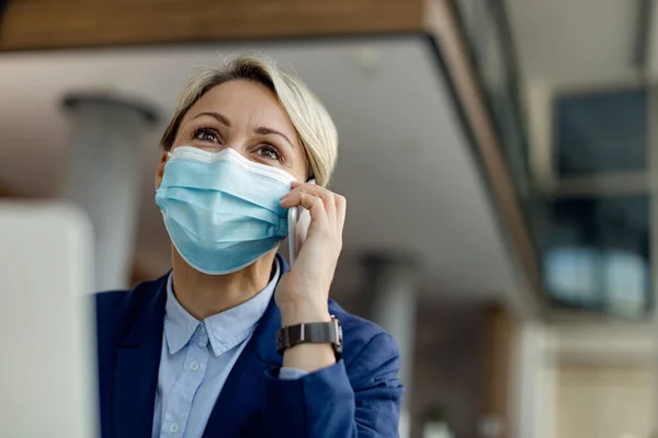 Low angle view of happy businesswoman communicating on mobile phone while working in the office and wearing face mask due to coronavirus pandemic.
