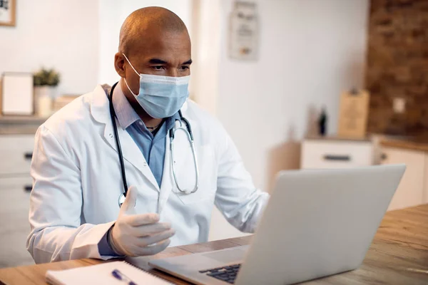 Black doctor wearing a face mask and consulting a patient via video call from his office due to COVID-19 pandemic.