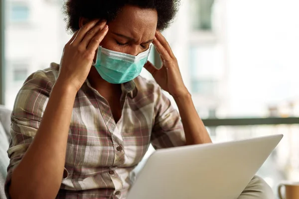 Exhausted black woman with face mask holding her head in pain while using computer at working at home.