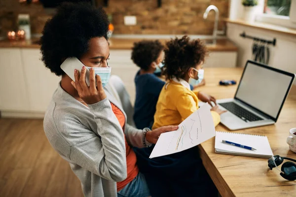 Black working mother talking on the phone and going through paperwork while her kids are using laptop beside her. They are wearing face masks due to COVID-19 pandemic.