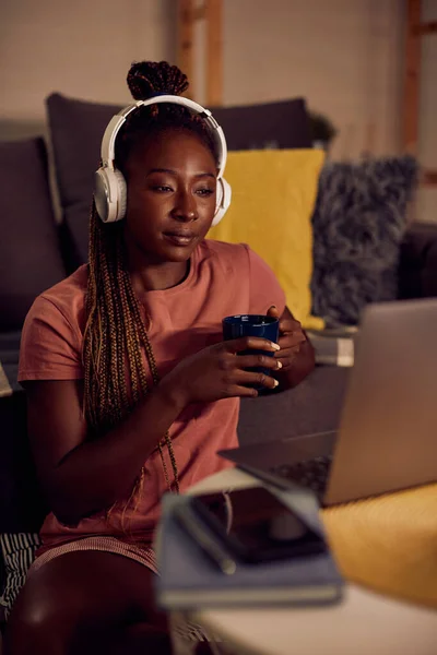 Young African American woman surfing the net on laptop while wearing headphones and having cup of tea in the evening at home.
