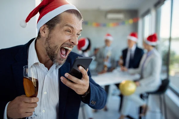 Male entrepreneur using mobile phone and screaming while drinking Champagne on New Year's party in the office. His colleagues are in the background.
