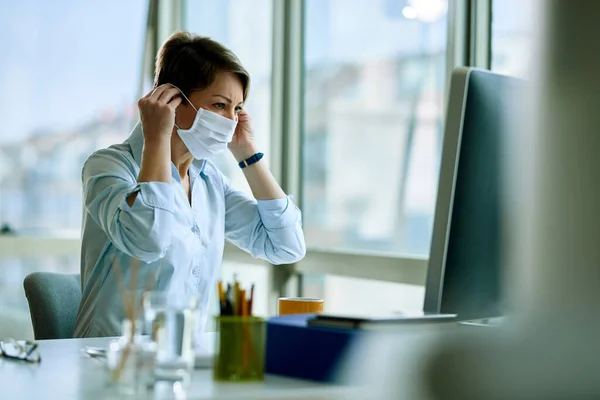 Businesswoman putting on face mask while working on a computer in the office during virus epidemic.