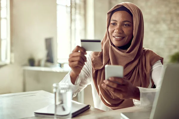 Smiling Muslim businesswoman shopping online while using credit card and mobile phone in the office.