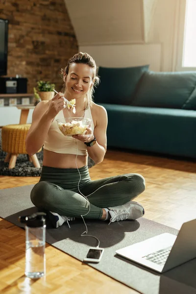 Happy athletic woman sitting on the floor and eating salad after exercising at home.