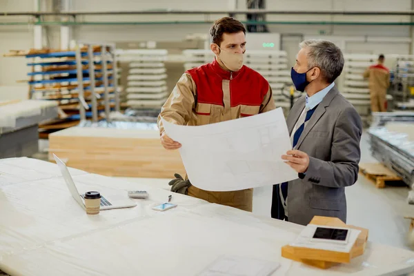 Company manager and and carpenter wearing protective face masks and cooperating while analyzing blueprints at industrial facility.