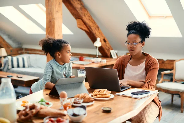 African American working mother using laptop while her daughter is using digital tablet during breakfast in dining room.