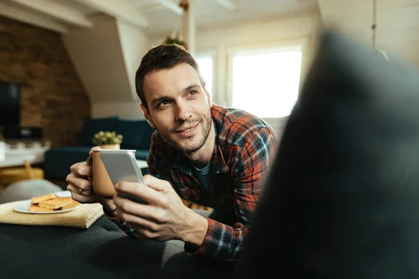 Young smiling man texting on mobile phone while relaxing in the living room and drinking coffee.