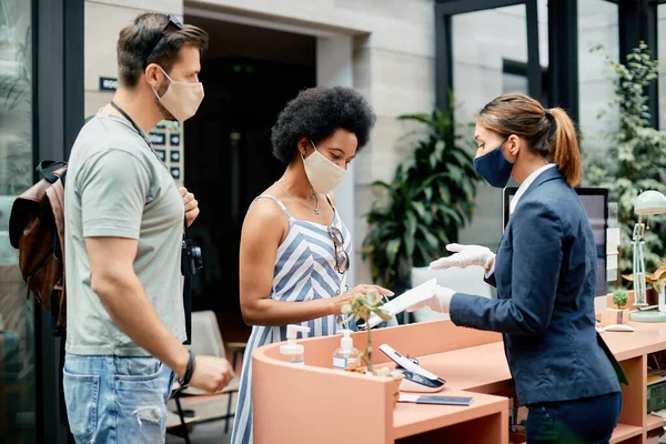 Female receptionist explaining application documents to a couple during their check-in a hotel. They are wearing protective face masks due to coronavirus pandemic.