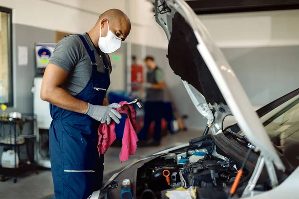 Black mechanic wearing face mask while repairing car\'s engine under the hood in a workshop.