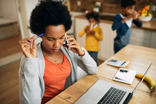 Black single mother talking on the phone while working on a computer at home. Her kids are in the background.