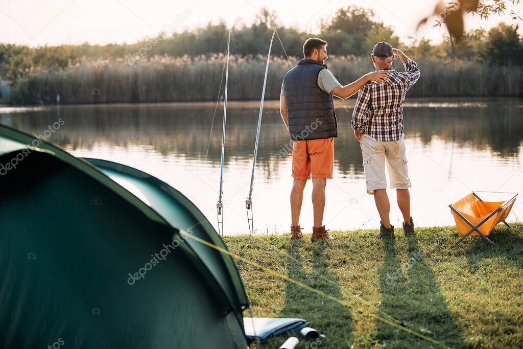 Back view of senior man and his son freshwater fishing on a lake while camping together.