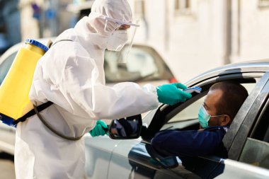 Healthcare worker in hazmat suit measuring temperature of a car driver with infrared thermometer at city checkpoint during coronavirus pandemic. 