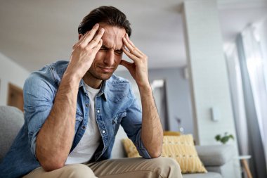 Low angle view of distraught man holding his head in pain while sitting in the living room. 