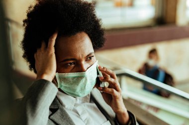 Distraught African American businesswoman with protective face mask making a phone call while being at airport corridor. 
