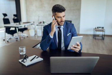 Young businessman working on digital tablet while talking on mobile phone in the office. 