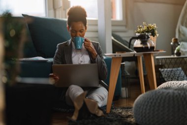Black female entrepreneur using laptop and drinking coffee while working at home. 