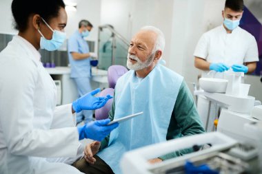 African American dentist using touchpad while communicating with senior patient during appointment and dentist's office. Focus is on senior man.