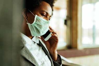 Worried African American businesswoman wearing face mask for virus protection while walking through public corridor and talking on the phone. 