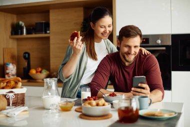 Happy man and his wife reading text message on cell phone during breakfast at home.