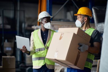 African American foreman with laptop communicating with male worker who is carrying carton boxes at distribution warehouse during coronavirus pandemic.