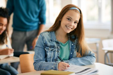 Happy high school student writing during a class at high school and looking at camera.