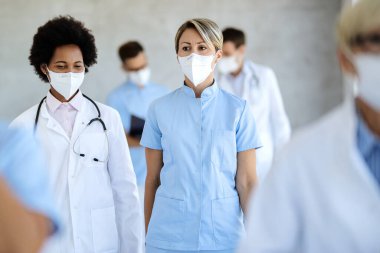 Young nurse and African American female doctor walking through hospital hallway with protective masks on their faces due to coronavirus pandemic. 