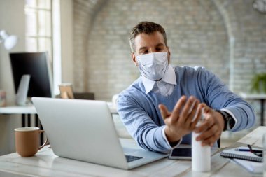 Businessman with protective face mask disinfecting his hands while working in the office. 