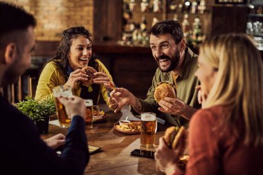 Young happy man having fun with is friends while eating hamburgers and drinking beer in a pub.