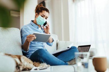 Young engineer with face mask communicating on cell phone while working on digital tablet at her home during virus epidemic. 