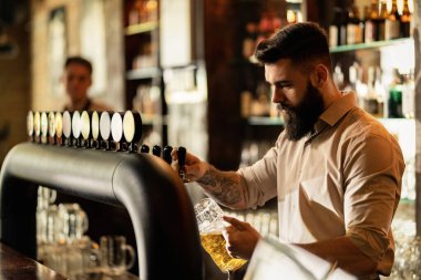 Young bartender pouring draft beer in beer glass while working at bar counter. 