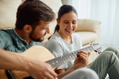 Young happy woman listening to her husband who is playing acoustic guitar at home.