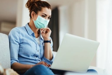 Young woman with face mask using computer and surfing the net while relaxing at home. 