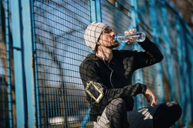 Tired male athlete drinking water form a bottle while resting after sports training outdoors. 